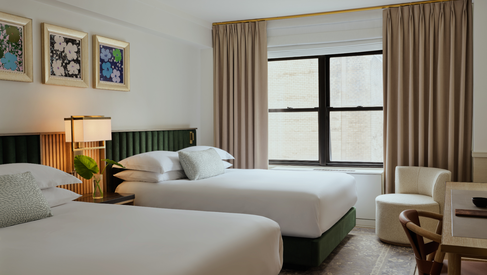A modern hotel room features two beds with green bases, pop art, and a desk near a large window